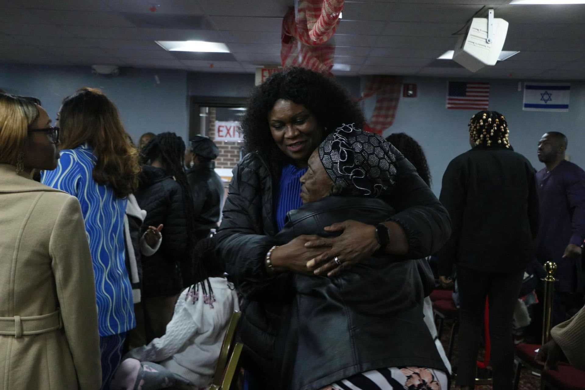 Two women embrace indoors, surrounded by people. One woman wears a black jacket, hugging the other.