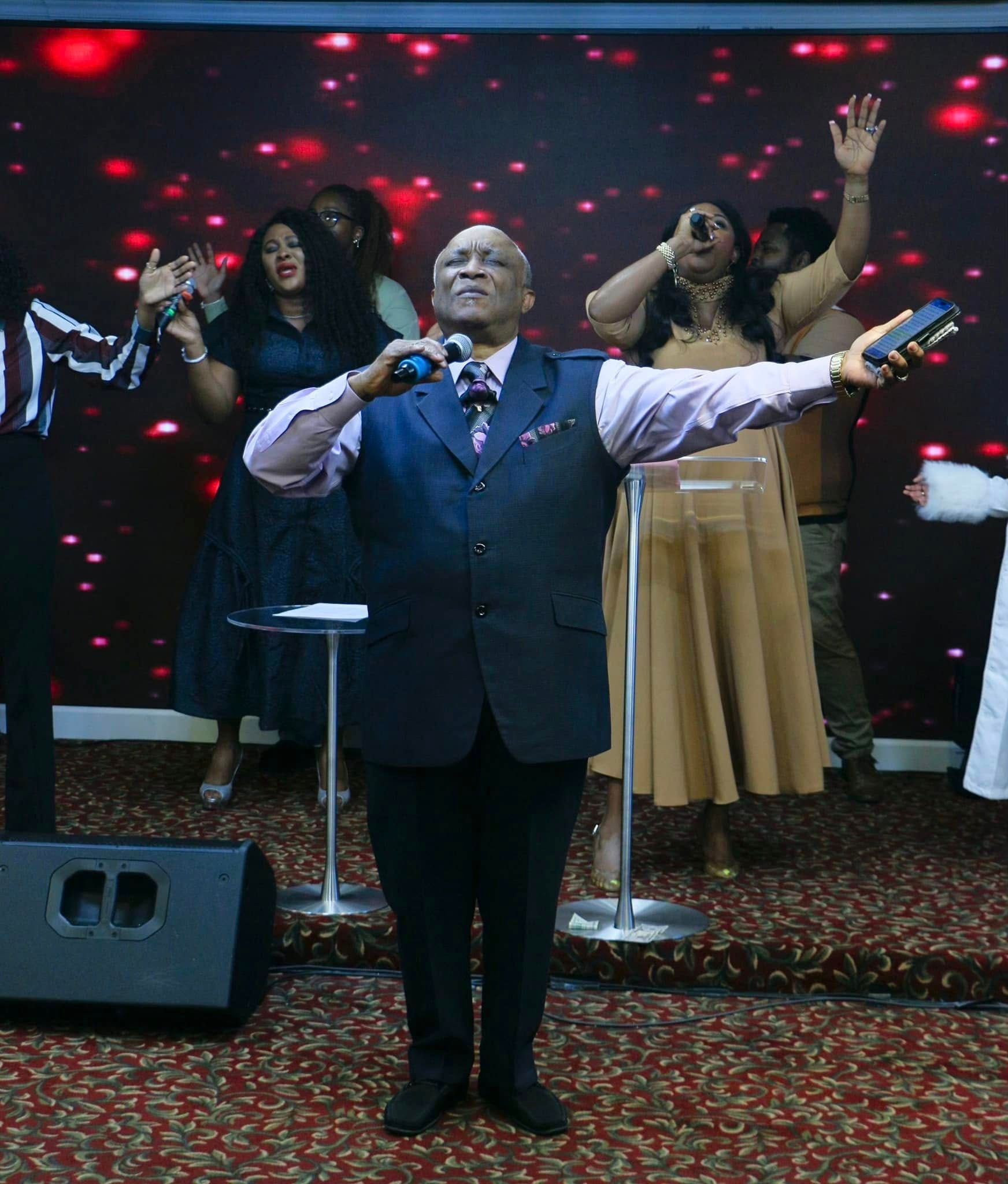 Man leading worship service with singers on stage, raising hands. Red background.