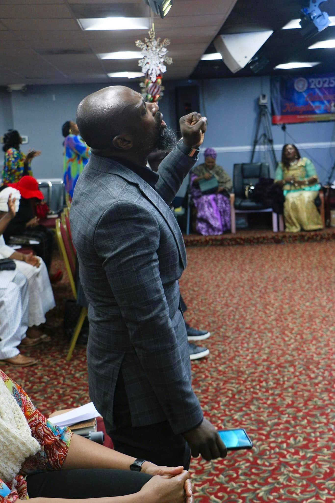 Man in gray suit raises arm, speaking to an audience. Indoors, room with people, decor, and stage.