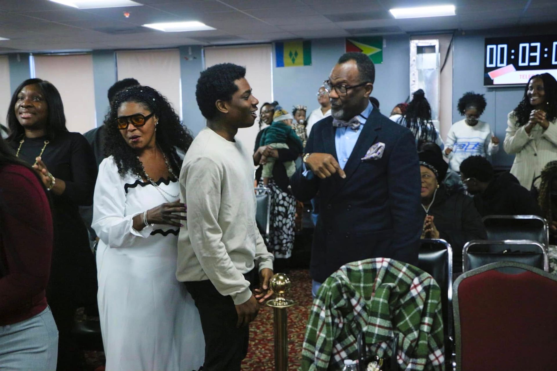 Group of people in a room. A man in a suit interacts with a younger man, others clap and smile.