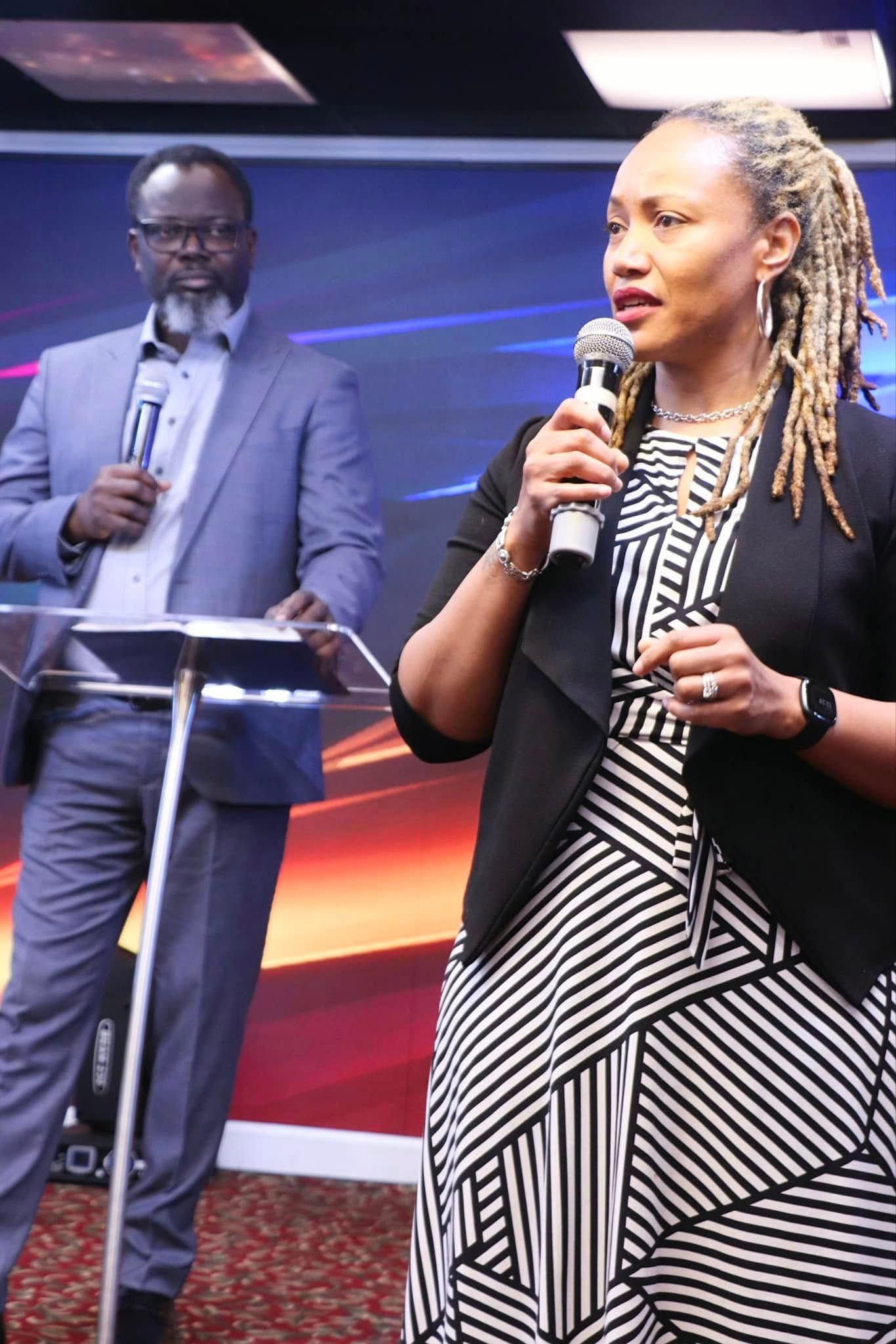 Woman speaks into microphone, man stands behind podium. Both are in a brightly lit room, possibly a church.