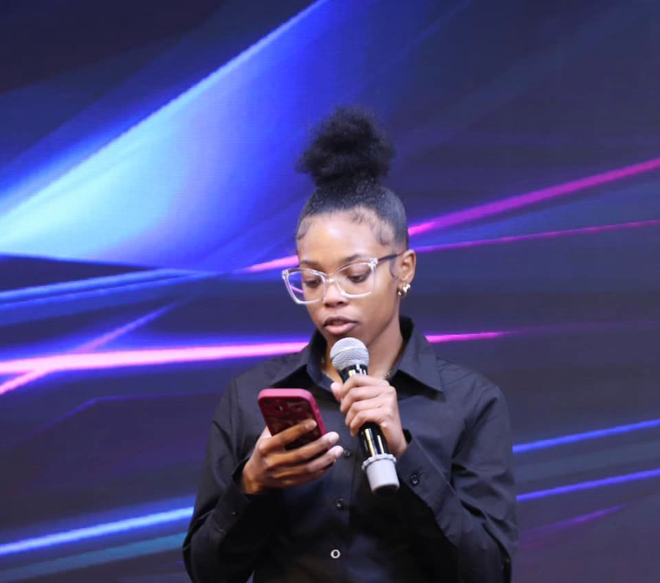 Young Black woman speaking into a microphone while looking at her phone on a stage with blue and pink lights.