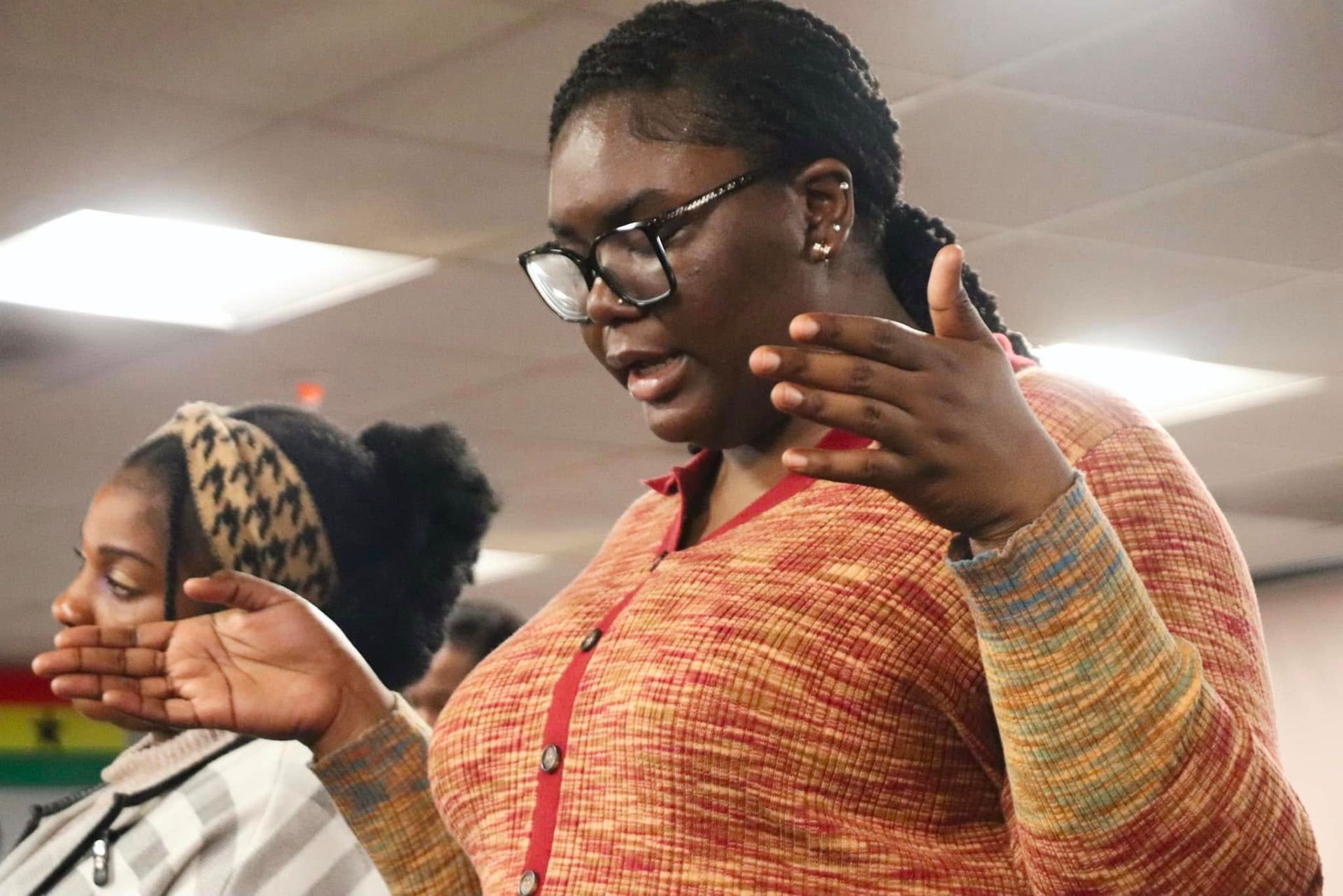 Woman with glasses speaks and gestures, indoors with others.