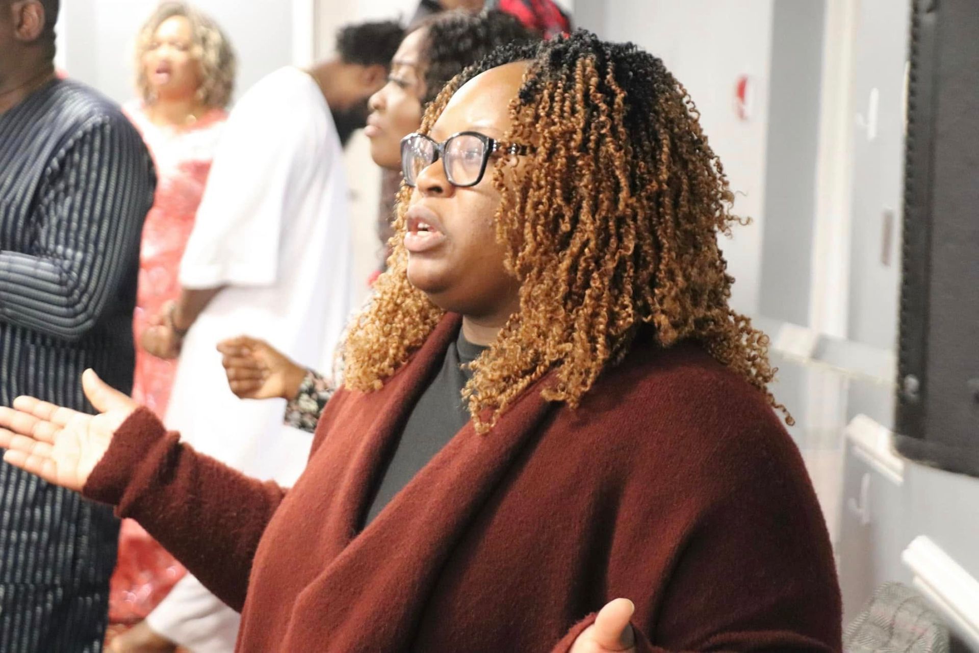 Woman with glasses and auburn hair, arms raised, singing in a church setting.