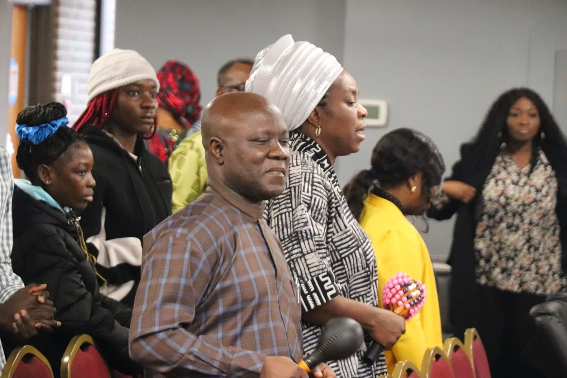 People in a room singing, some wearing head wraps, one man in a patterned shirt.