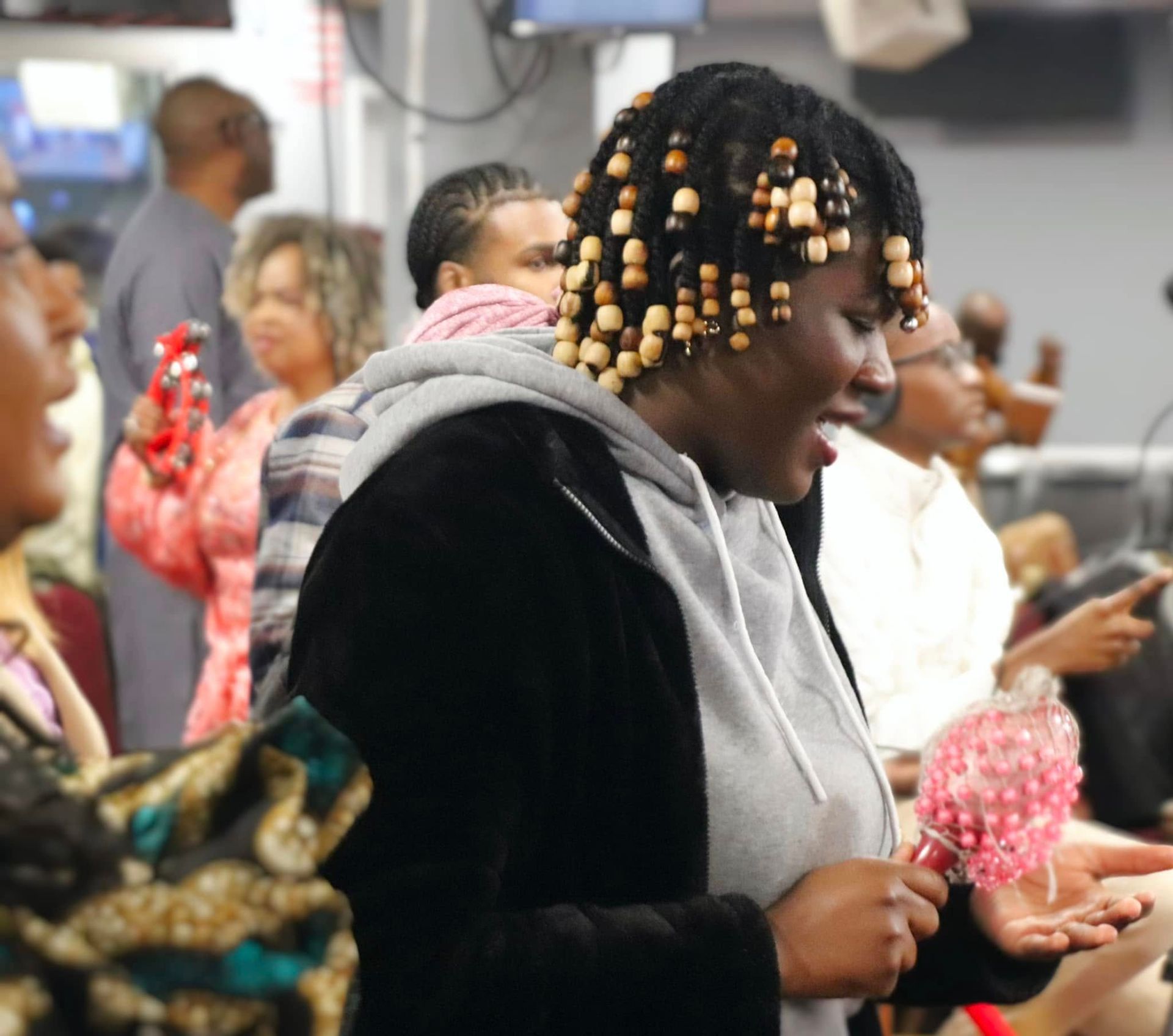 Woman with beaded hair sings, holding a shaker, smiling in a crowded room.
