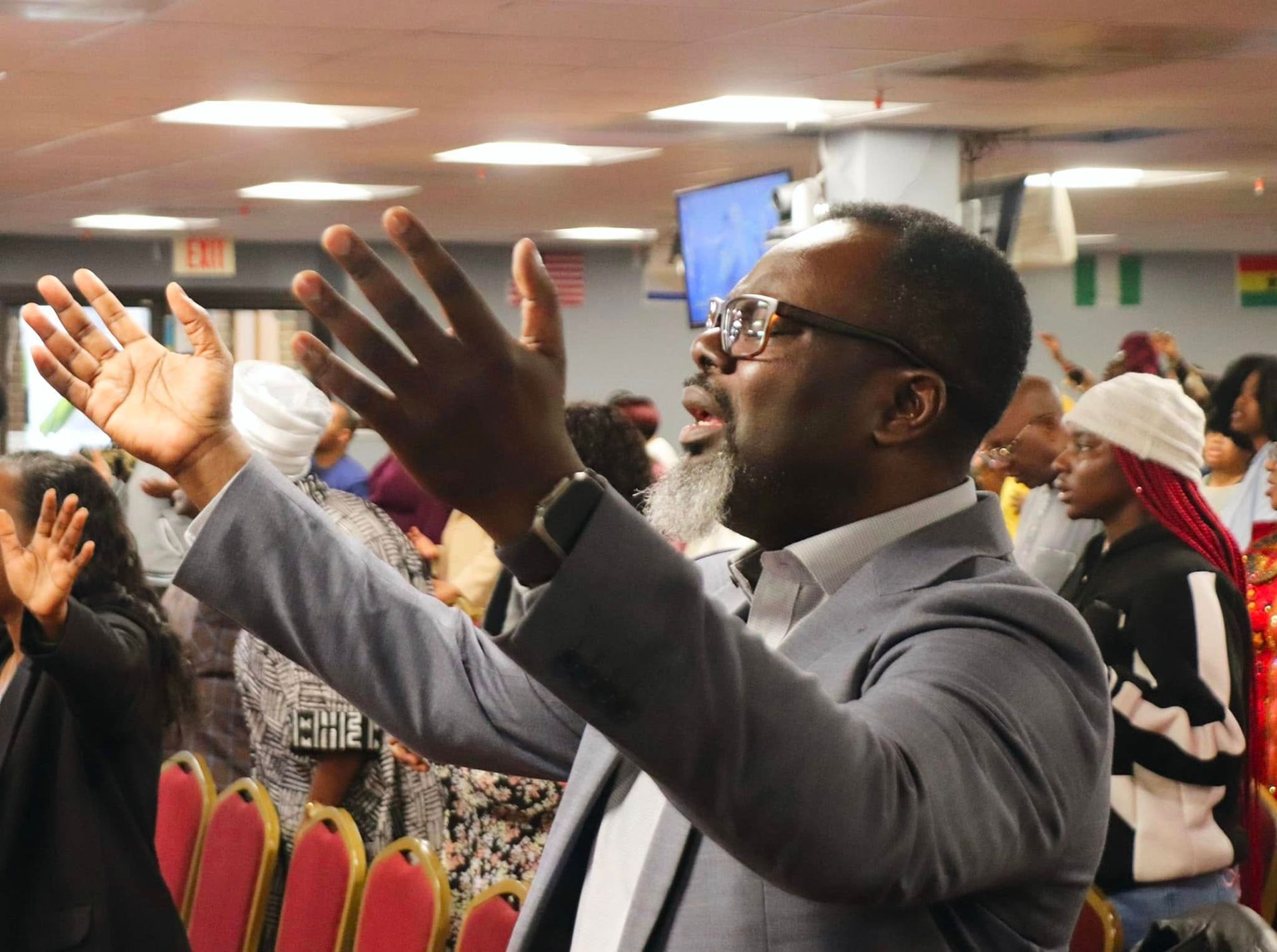 Man in suit raises arms in worship, eyes closed, with others in a church setting.