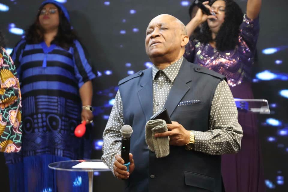 Man in suit leading a worship service, holding a microphone and cloth; women sing behind him.
