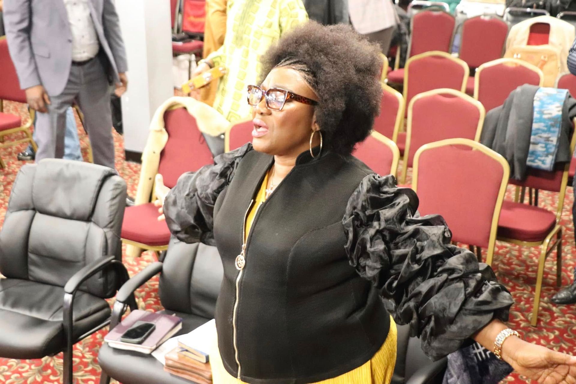 Woman in black vest with ruffle sleeves speaks, in room with chairs.