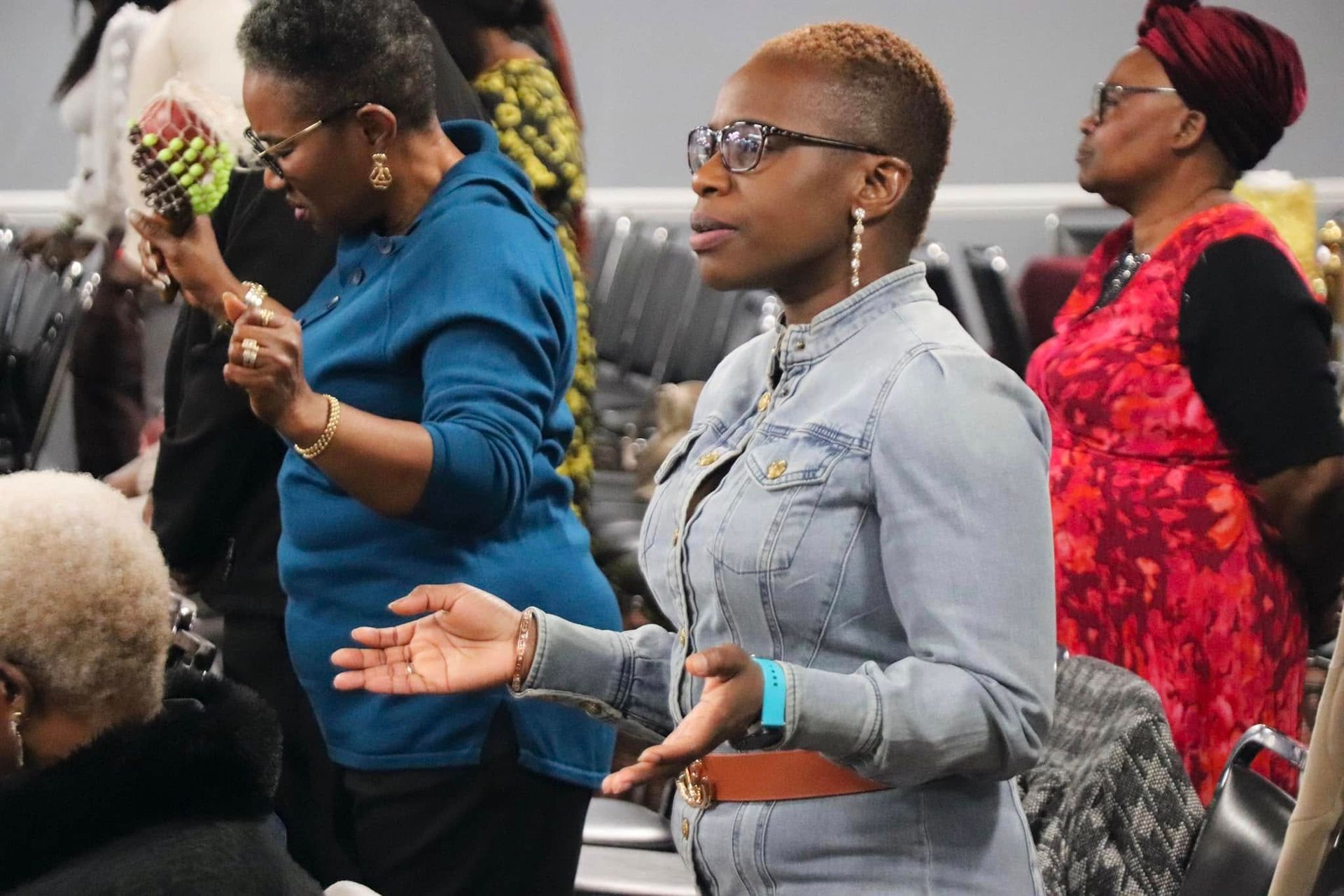 Woman with short hair praying in a group; arms raised, wearing a denim jacket.