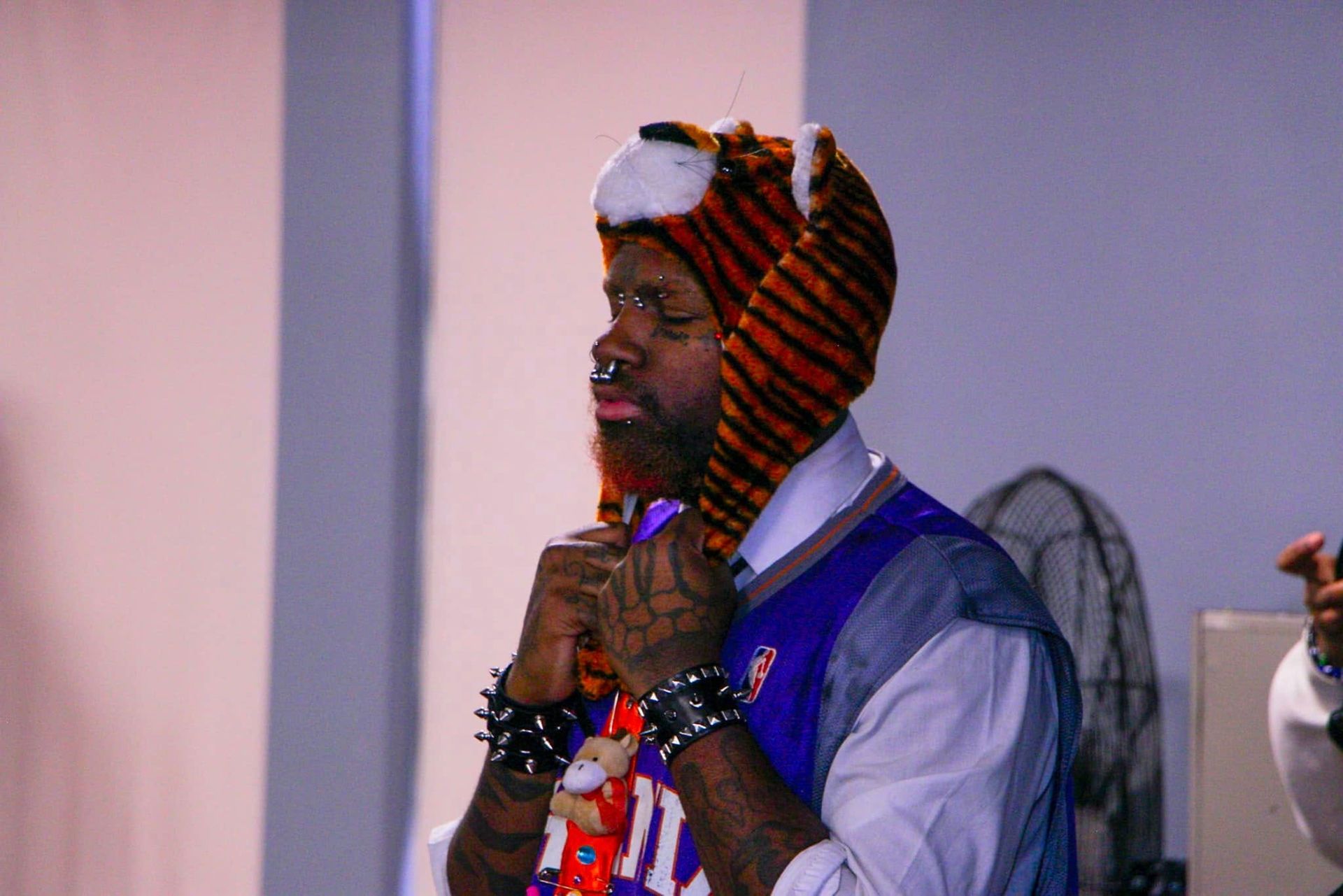 Man in tiger hat, adjusting it. Wearing spiked wristbands, purple vest over white shirt, indoor setting.