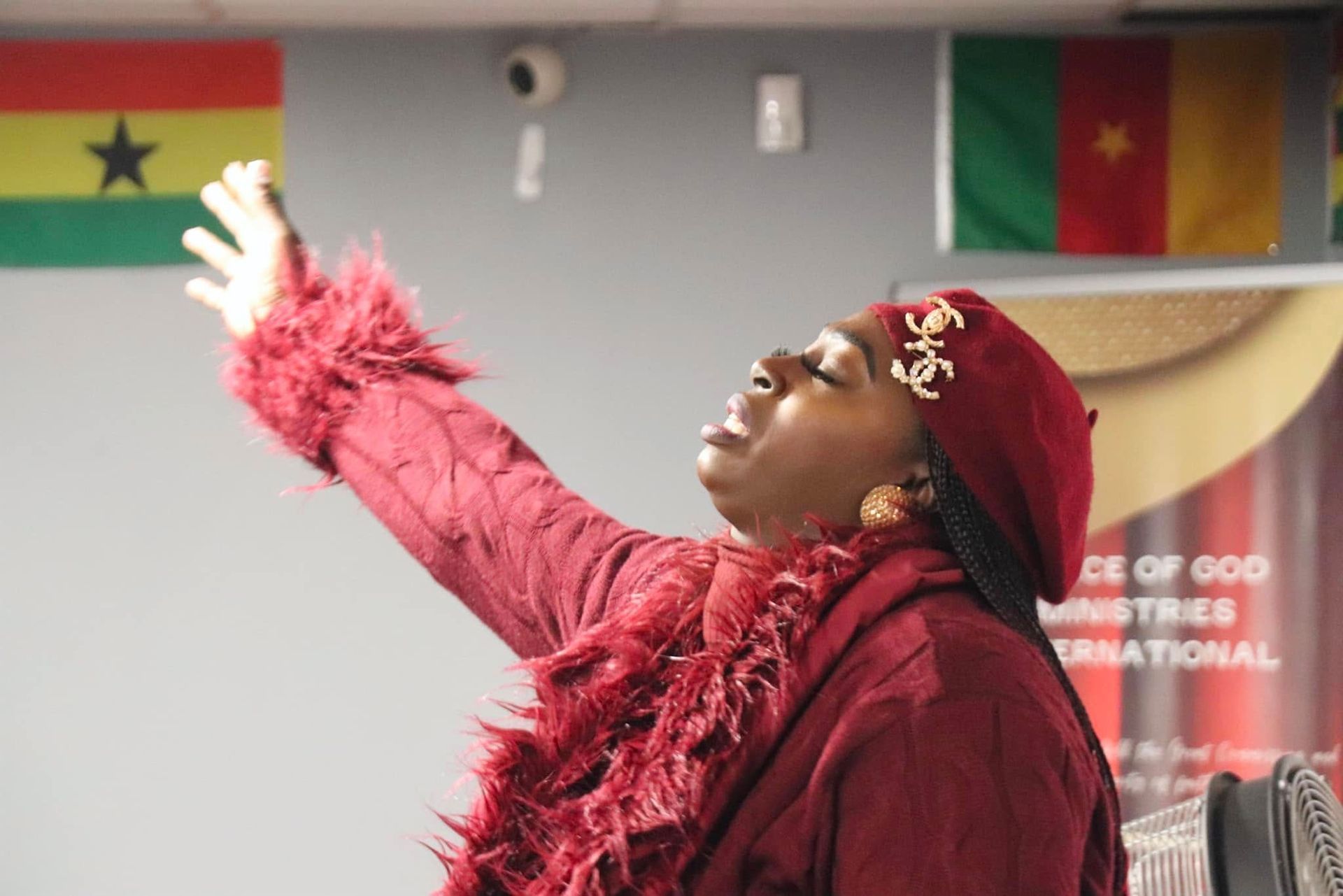 Woman in maroon, arms raised in praise, eyes closed, flags of Ghana and Cameroon in background.