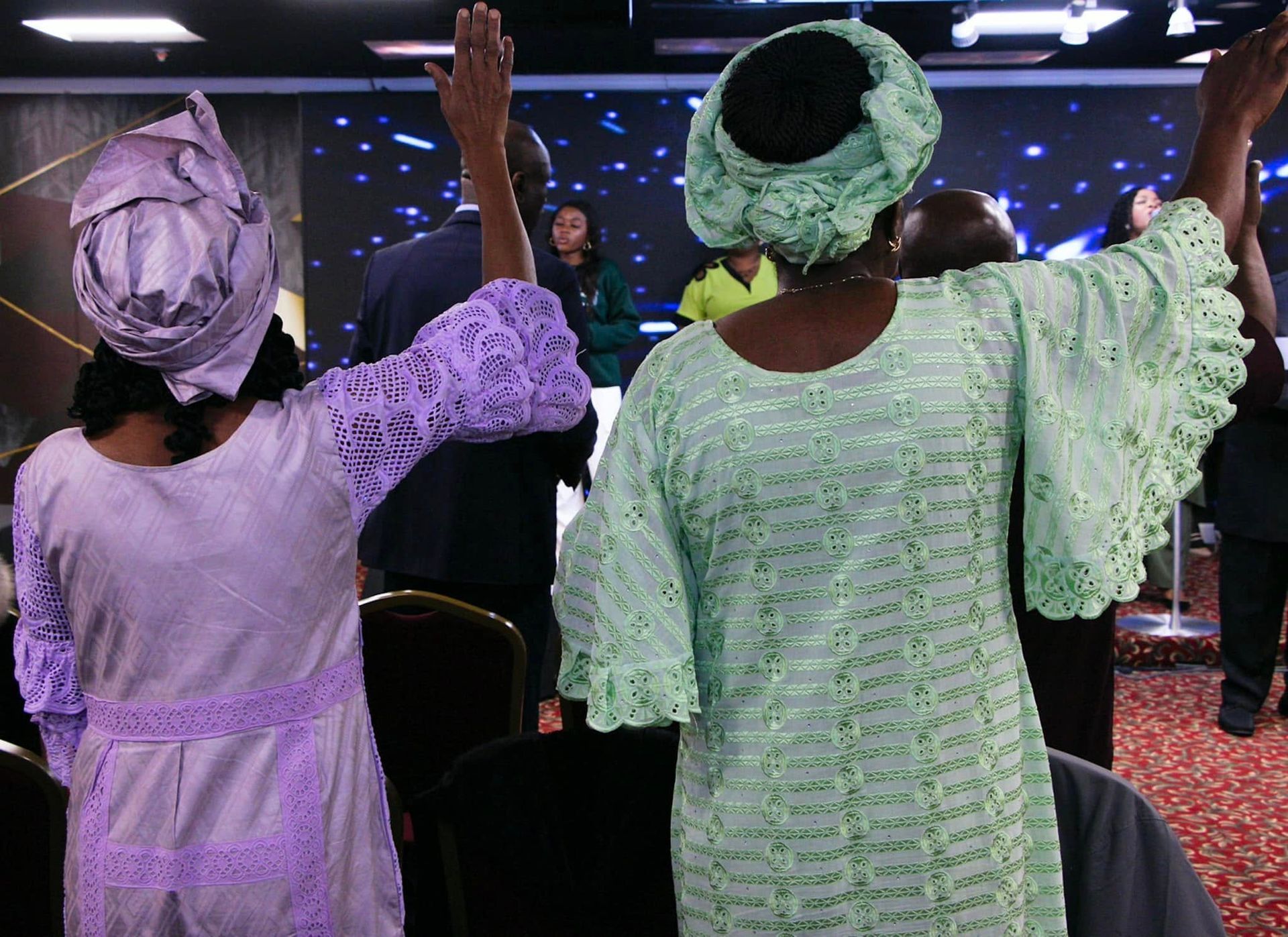 Two women in church, arms raised in worship. One wears lavender, the other mint green, both with matching head wraps.