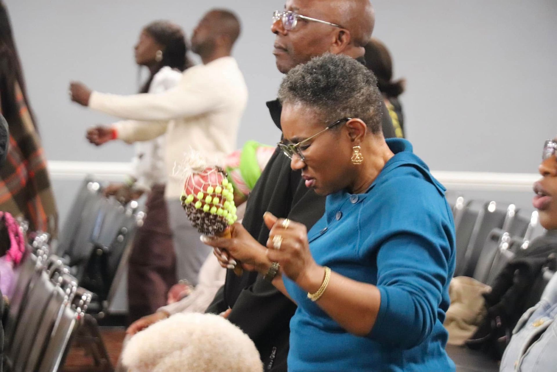 People in a room; woman in blue top playing a maraca, others raising hands in worship.