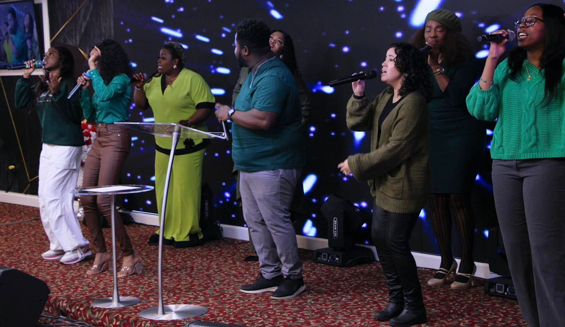 Choir singing on stage with a starry backdrop. People in various green, brown, and black outfits.