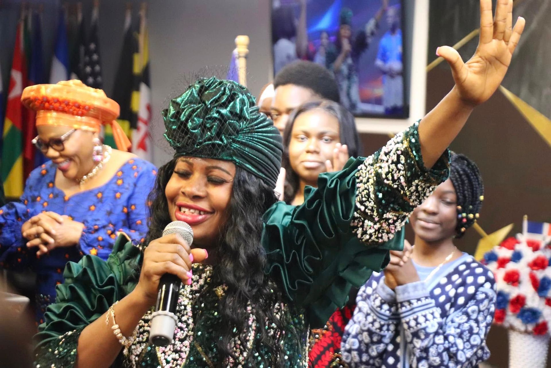Woman in green singing with hand raised, others in background. Stage setting, flags, people watching.