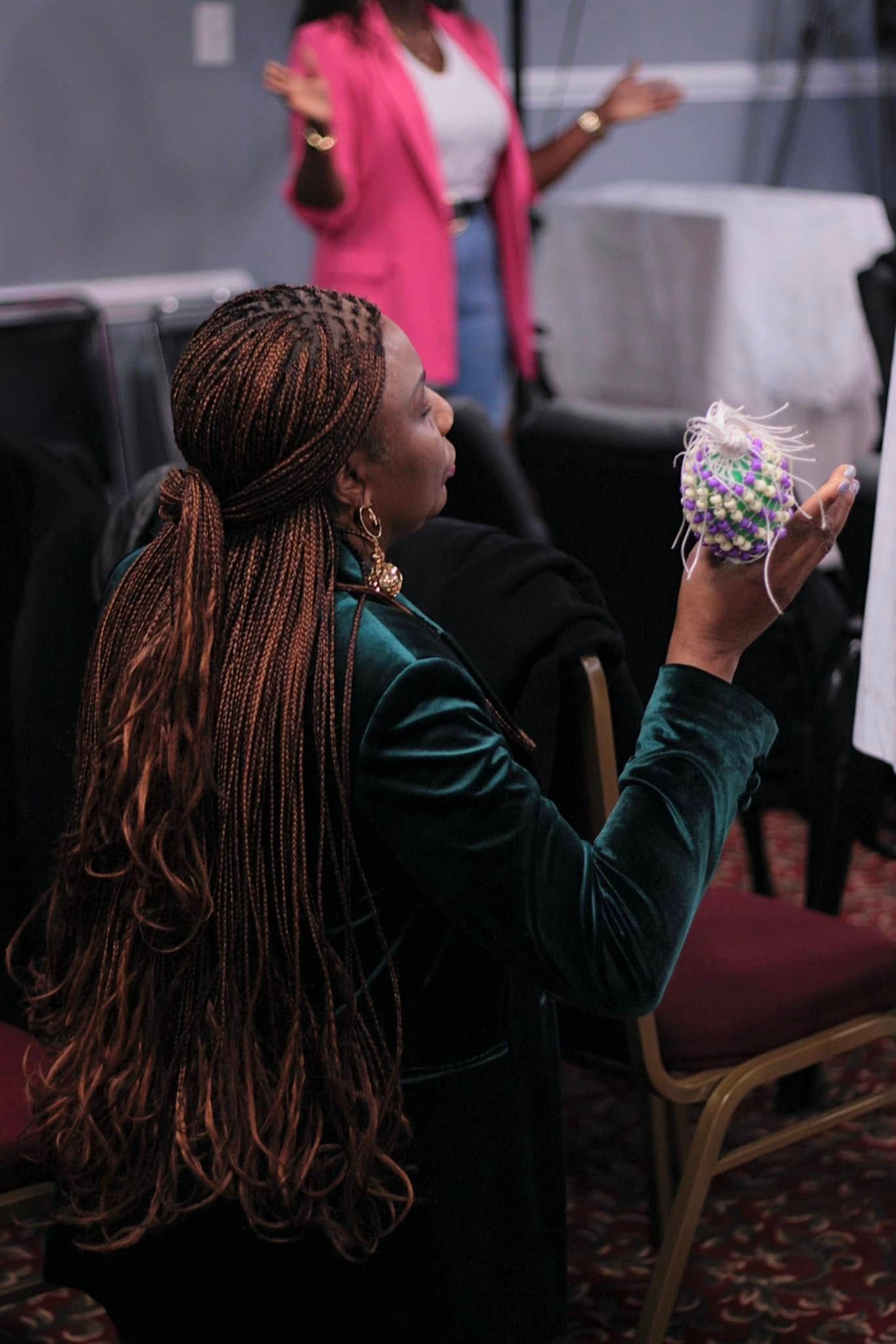 Woman with long braids in green velvet jacket holding object; another woman in pink blazer stands.