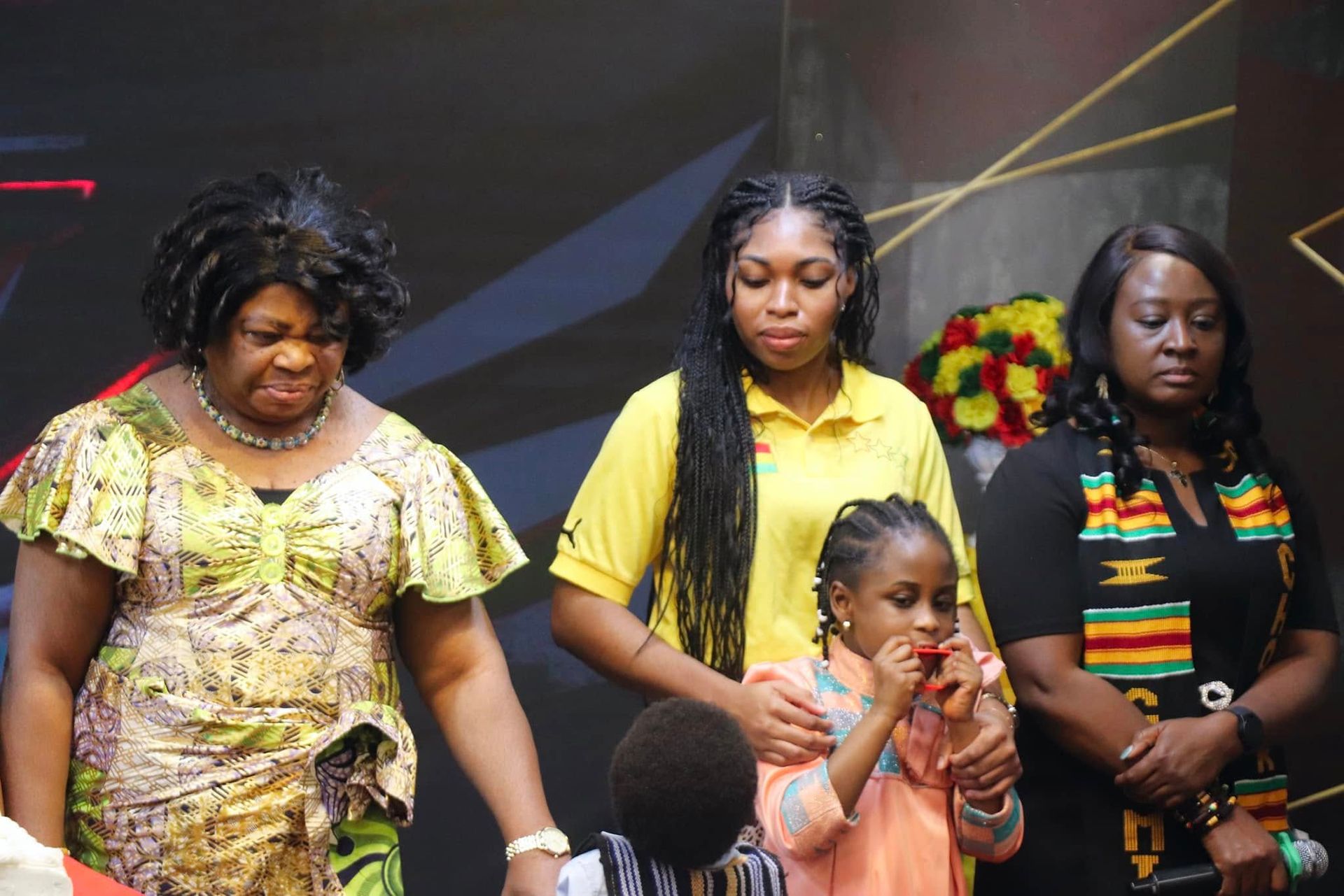 Three women and a child stand onstage, one blowing a noisemaker.
