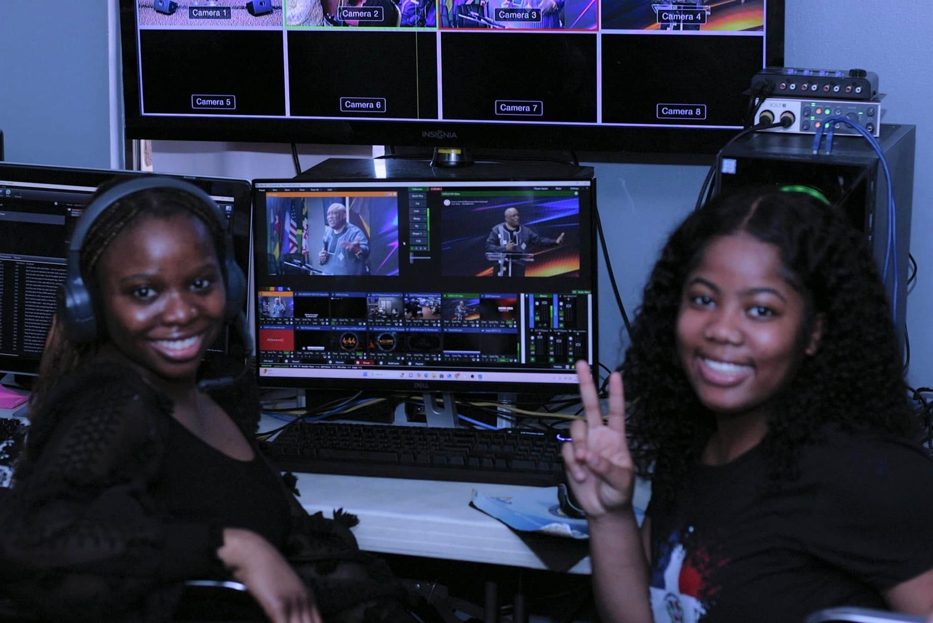 Two young women smile at the camera in a control room, surrounded by monitors and tech equipment.
