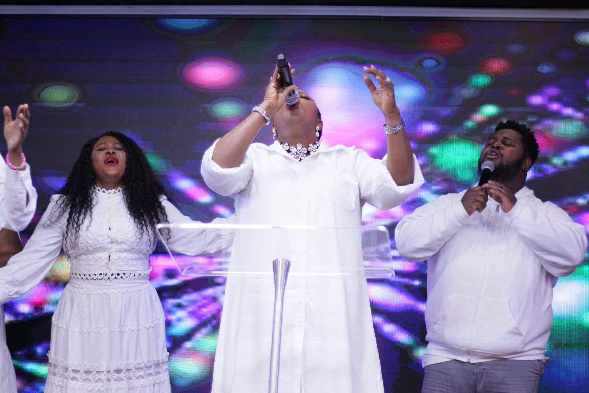 Choir singing on stage, dressed in white. Woman singing with raised arms, others singing with open hands.