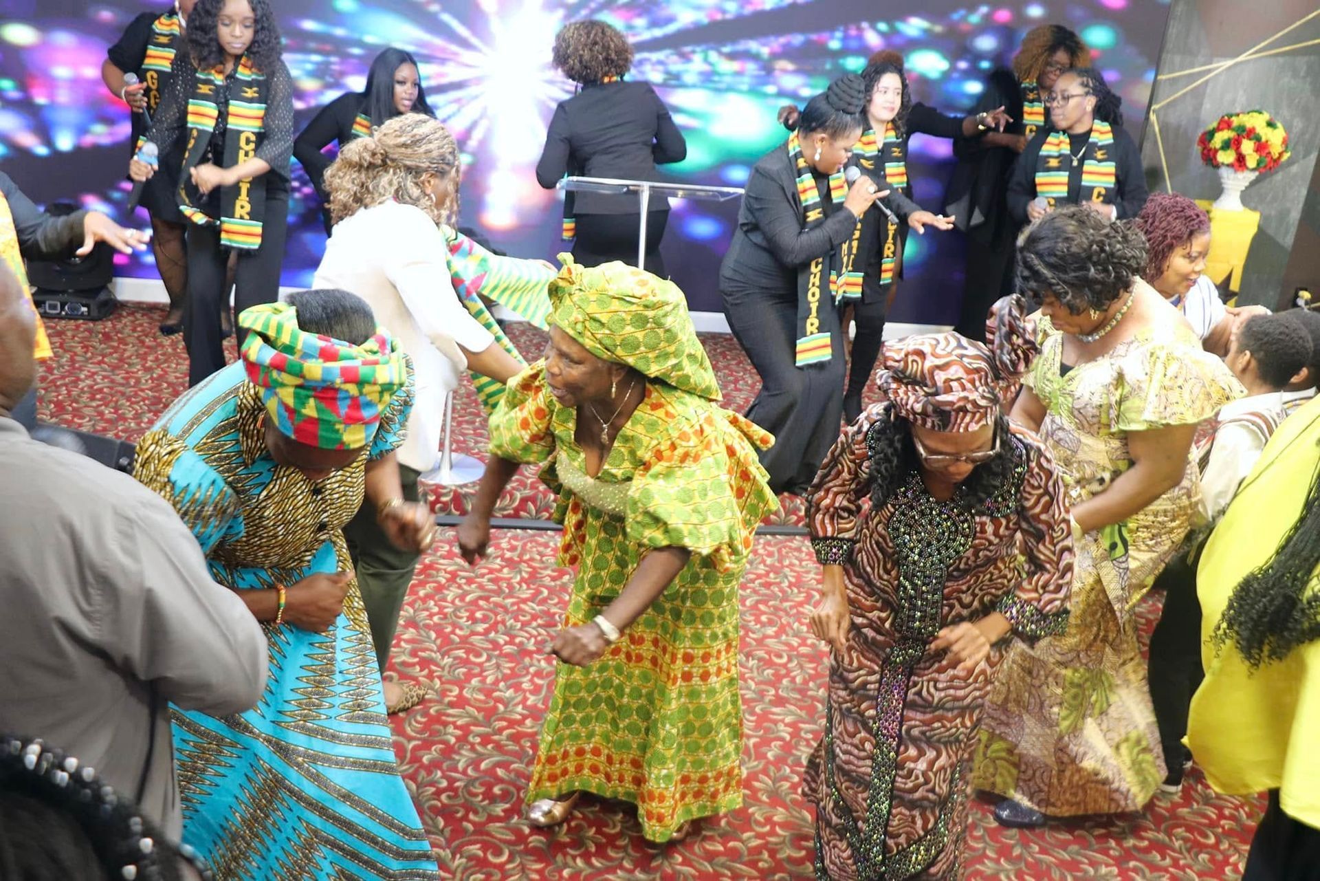 People dancing in colorful African attire at a gathering, indoors with a stage backdrop.