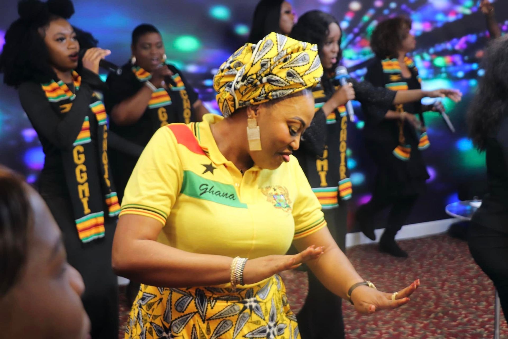 Woman in yellow shirt and headwrap dancing with a group wearing black outfits; background with lights.