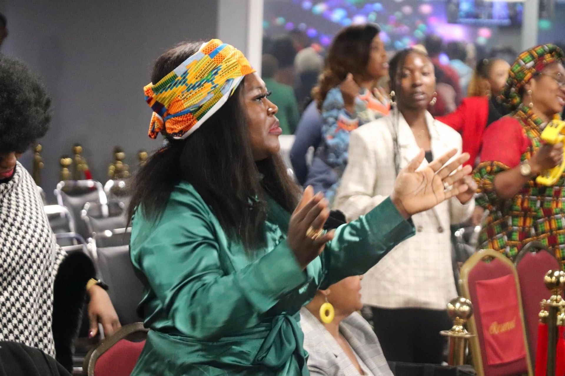 Woman in green robe with hands raised, worshiping in a church. Other people are in the background.