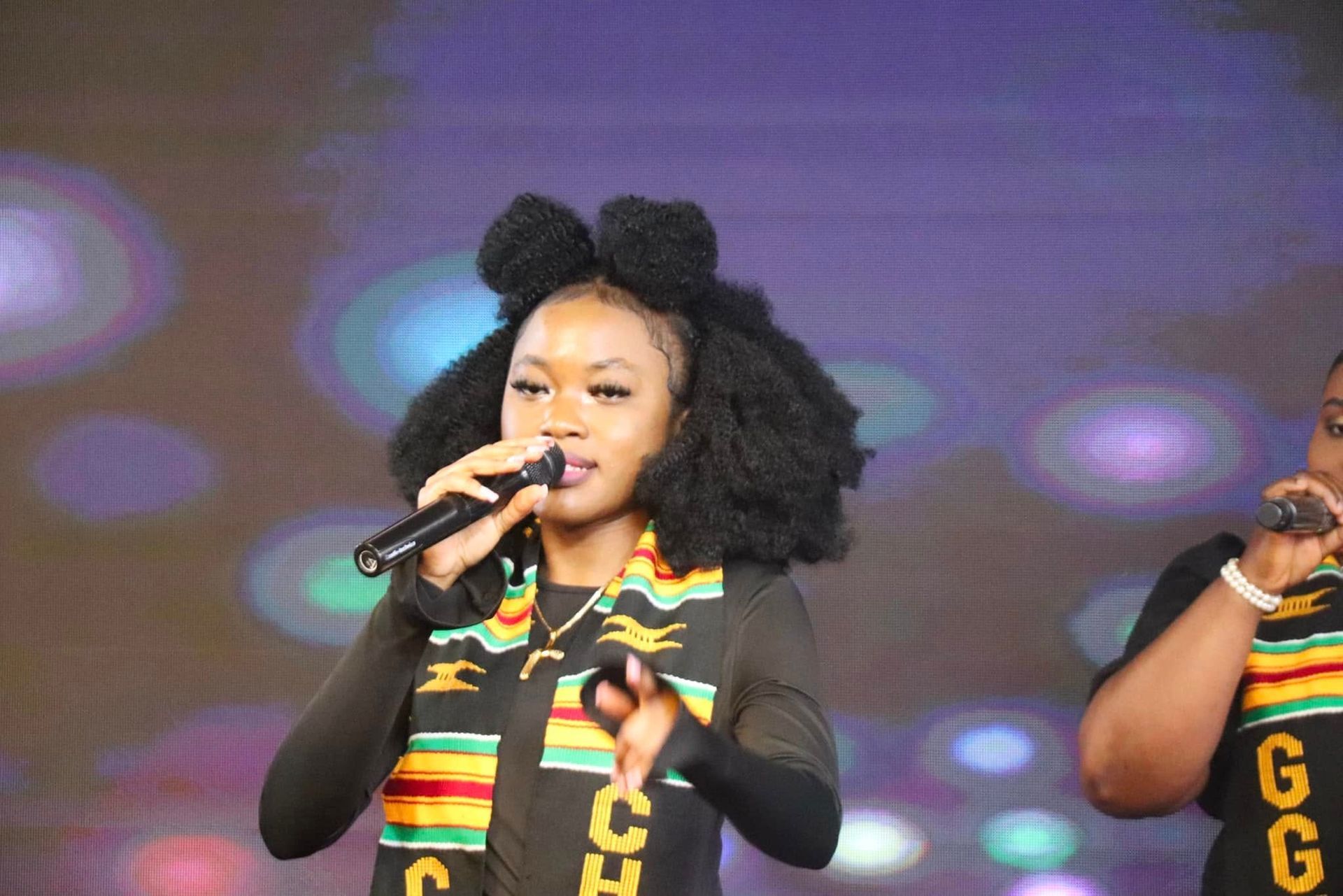 Young Black woman singing into a microphone onstage, wearing a graduation stole and hairstyle with bows.