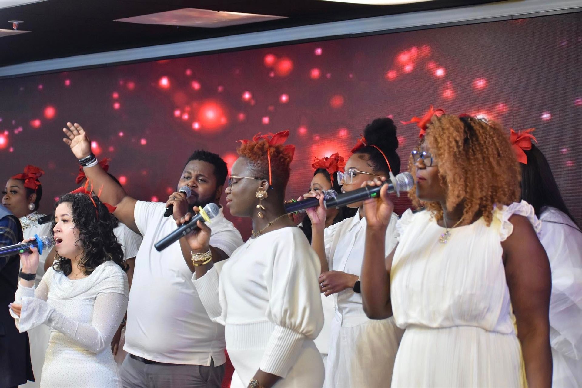 Choir singing with microphones on stage. White outfits, red accents, red and white bokeh background.