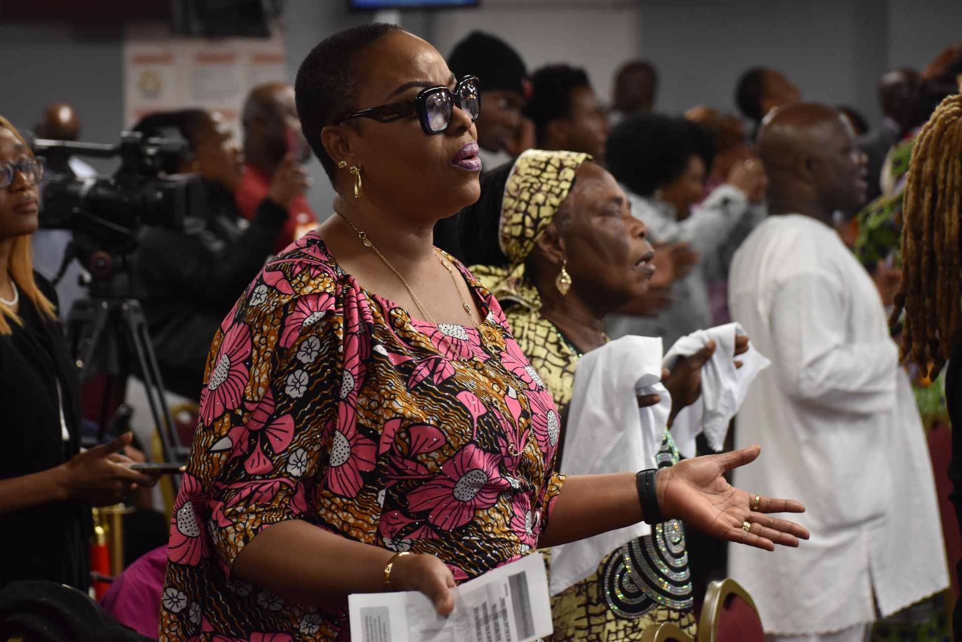 Woman in patterned top, sunglasses, and raised arms, praying in a church.