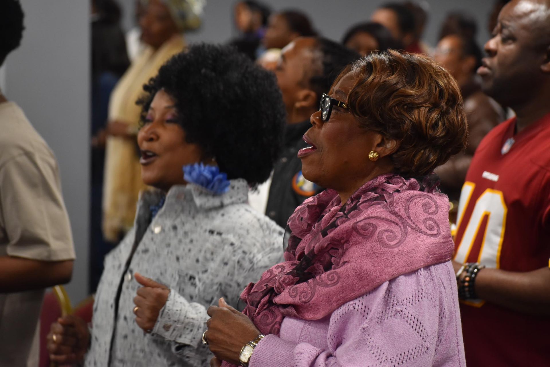 Two Black women singing enthusiastically, heads tilted upward, at an event with a crowd.