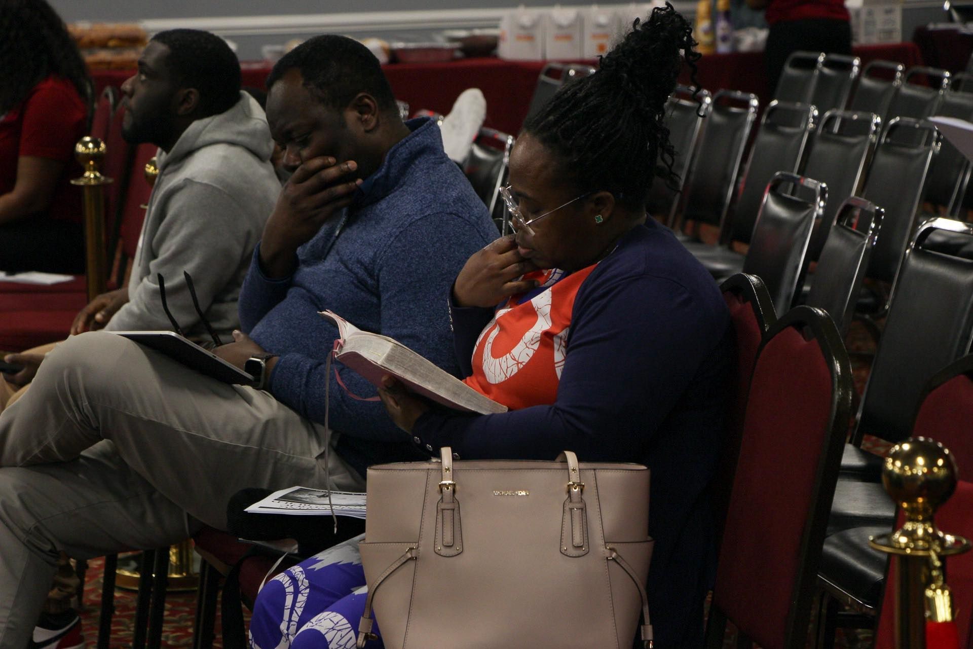 People in a church reading, listening, and attending a service. Some are looking at books, seated in rows.