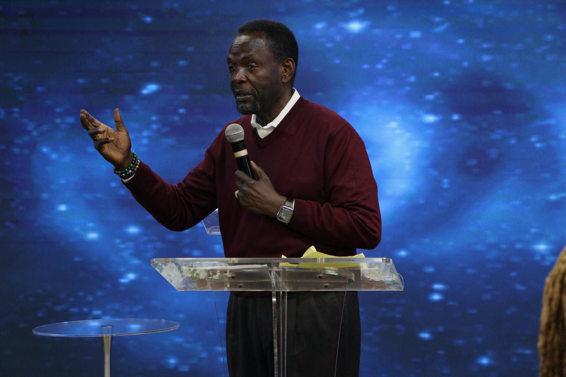 Man in maroon sweater speaking at a lectern, blue background.