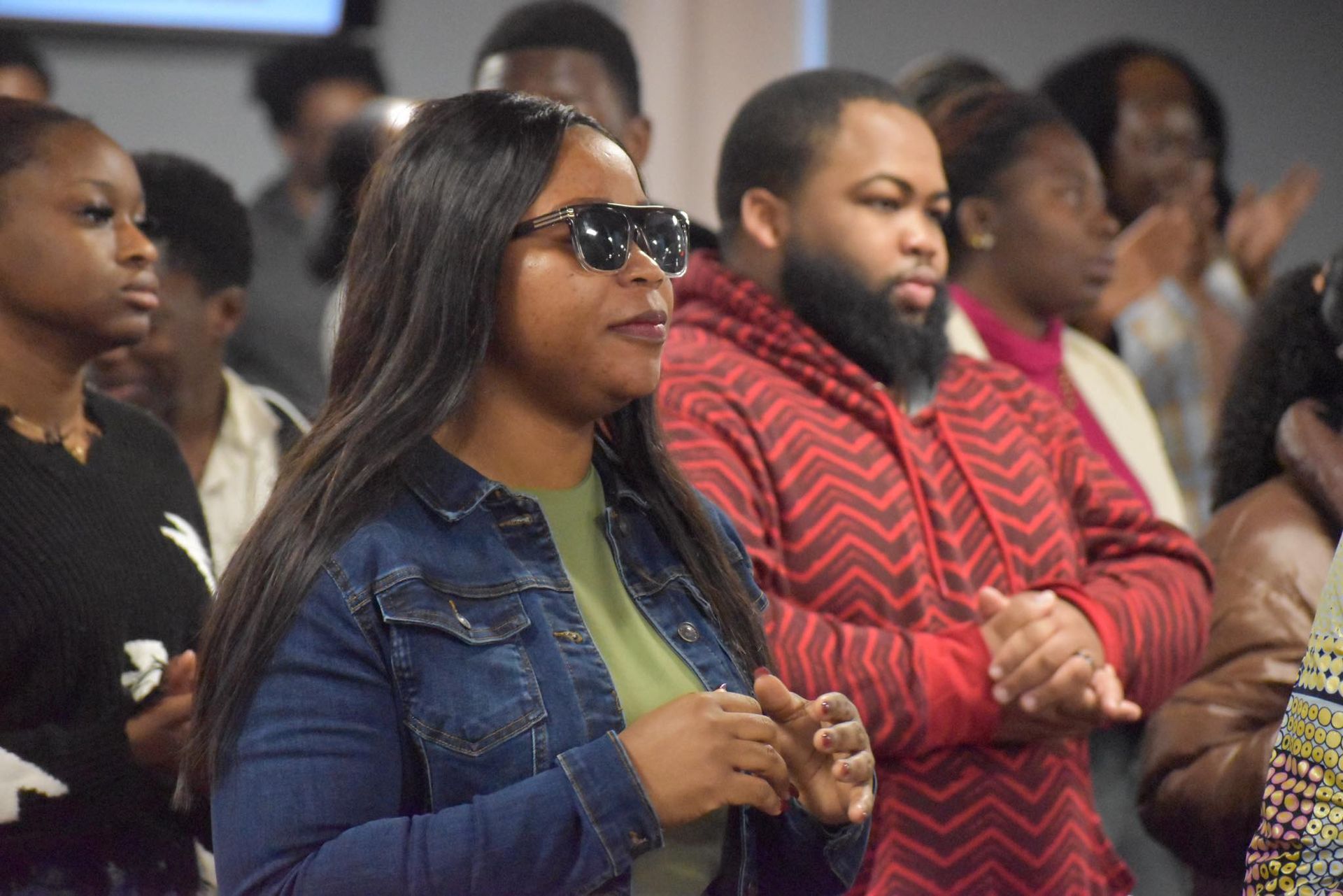 Group of people in a room, some with hands clasped, listening. Woman in sunglasses, jean jacket, and green shirt.
