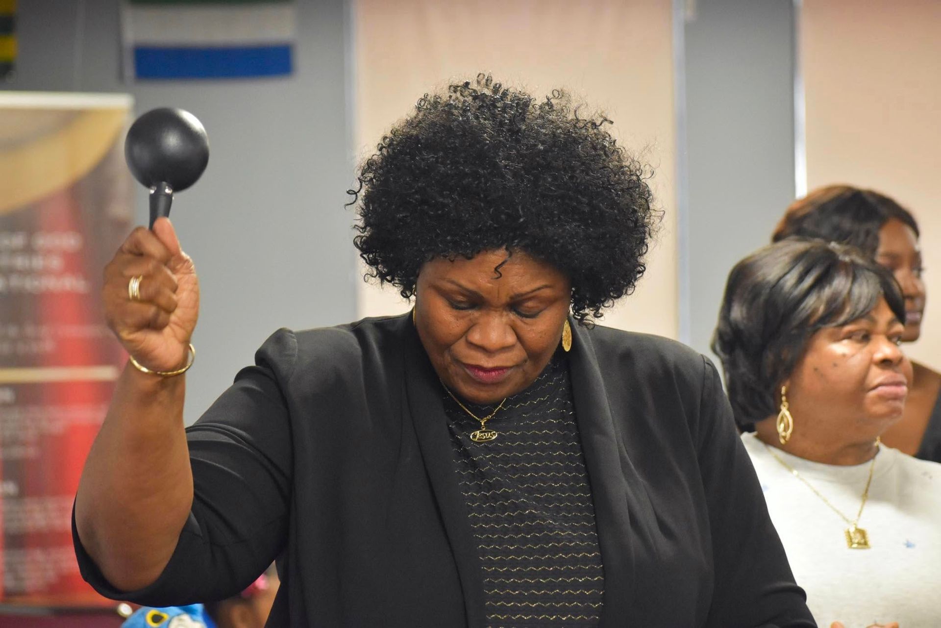 Woman in black blazer holds a black object up, eyes closed. Other women in the background.