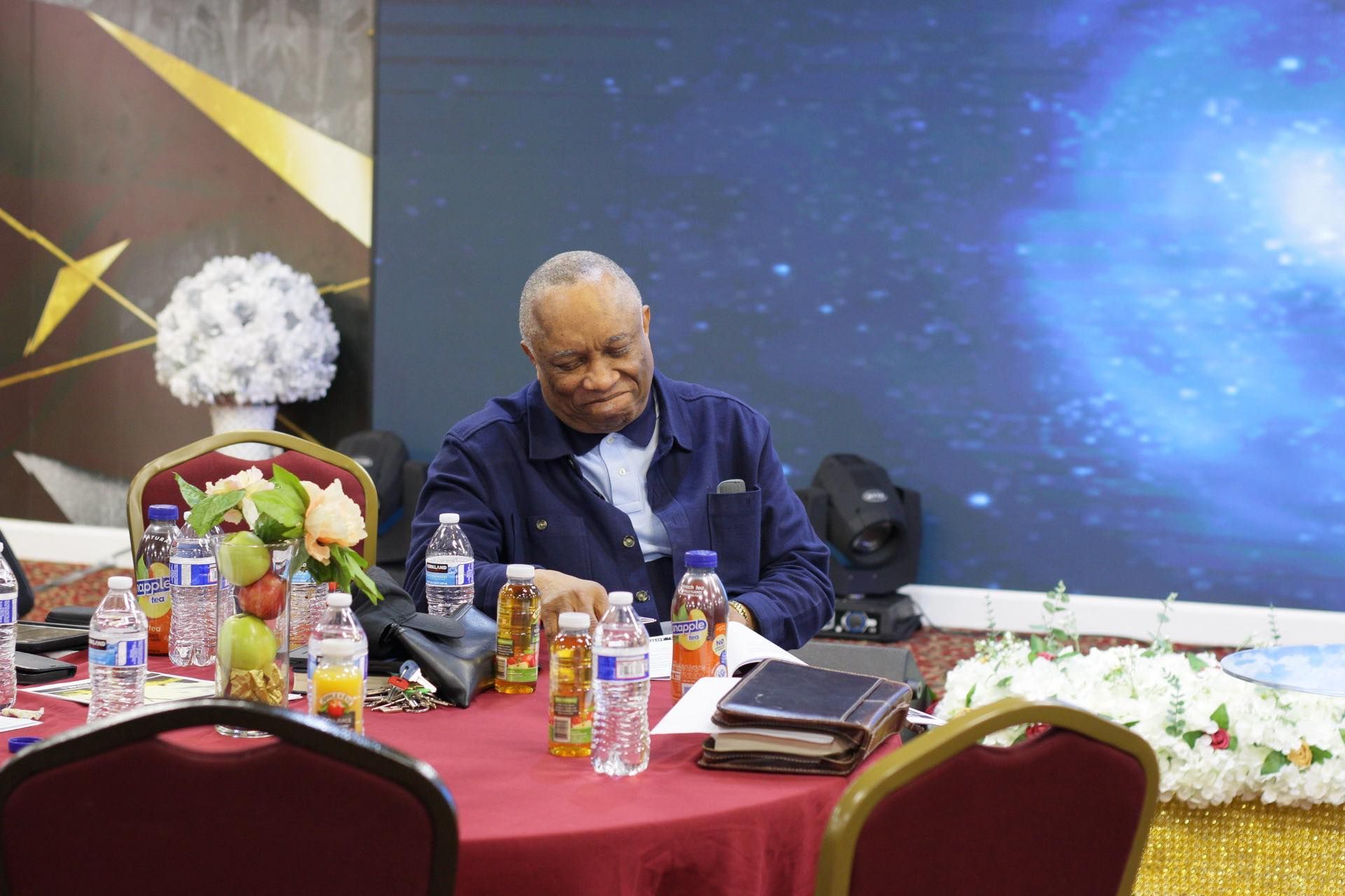 Man at table with drinks, papers, and flowers. Blue background, red tablecloth.