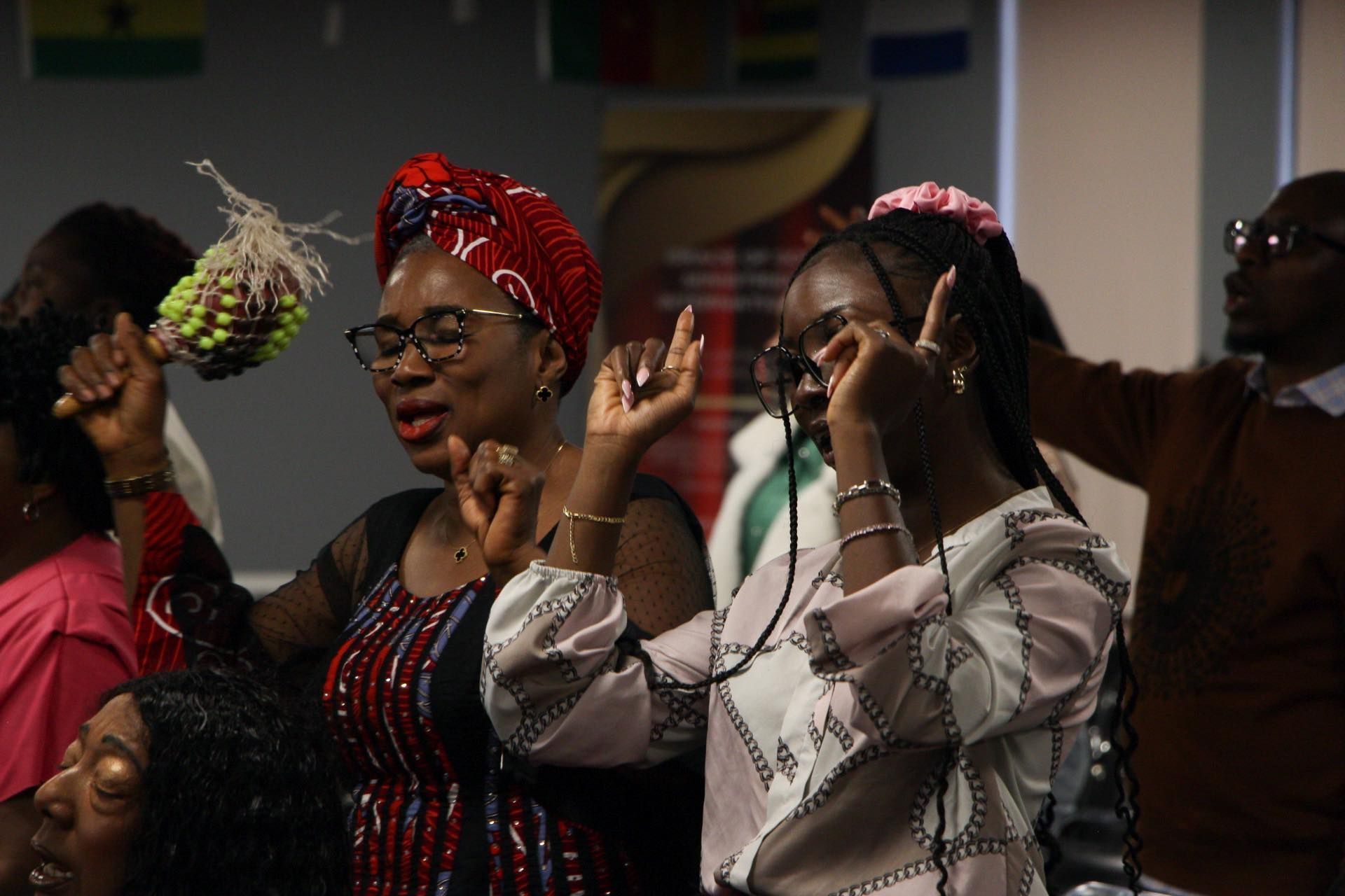 People in joyful worship, raising hands, and singing. One woman holds a maraca. Indoors, flags in background.