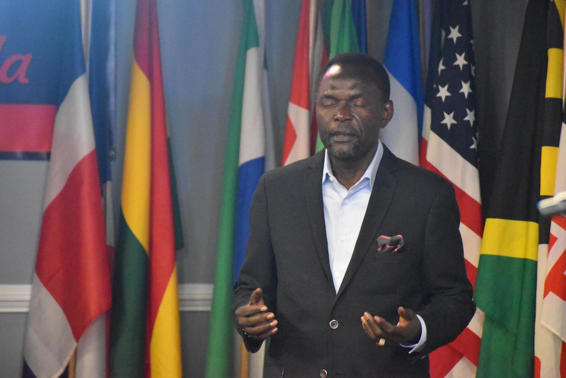 Man in a suit speaking, hands up, in front of flags of various nations.