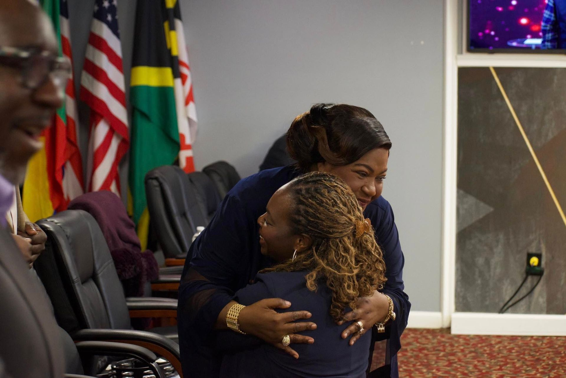 Two women in formal wear embrace, others in a room with flags.