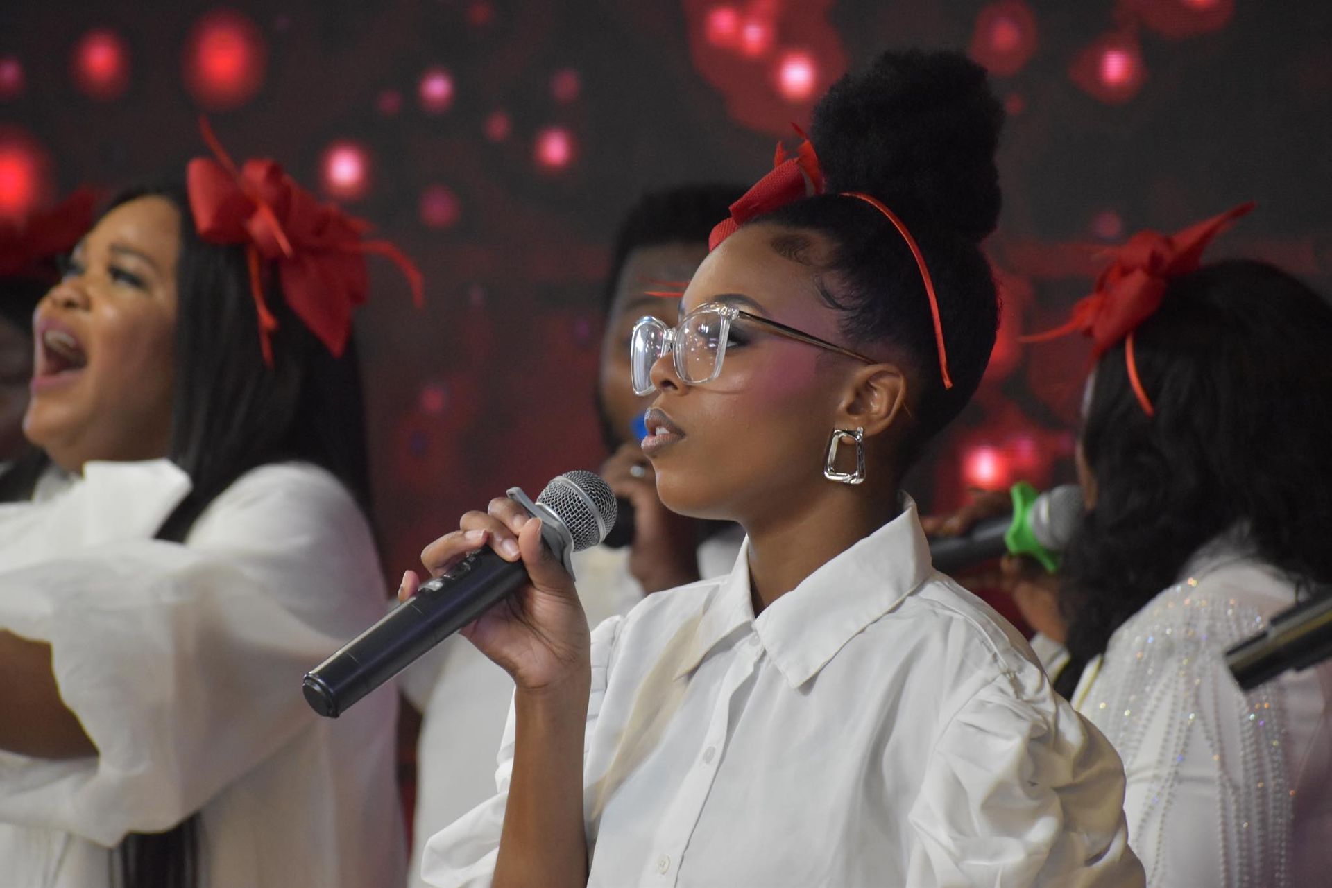 Choir of people wearing white singing, red background. Woman with glasses sings into a microphone.