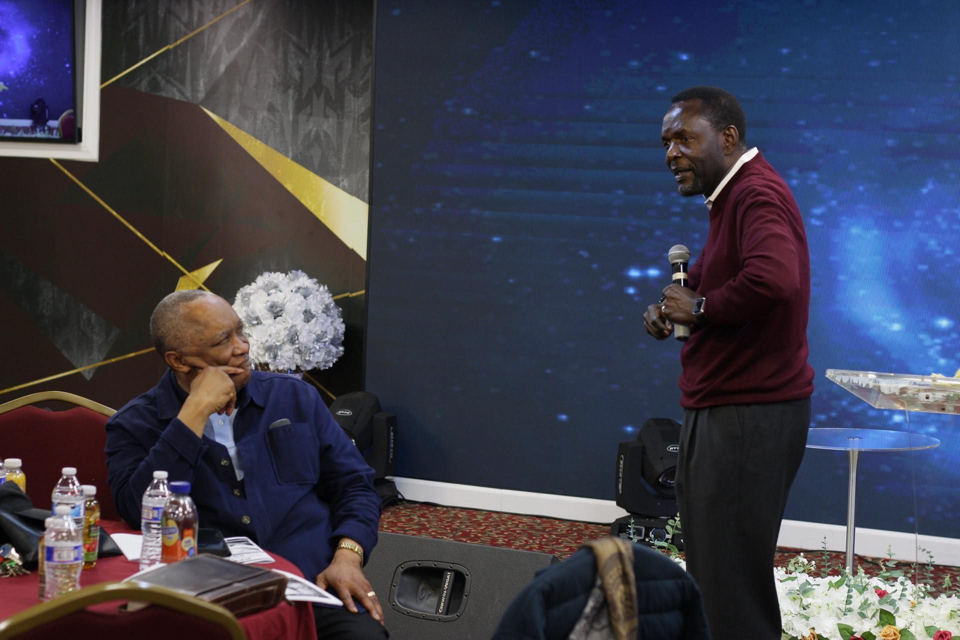 Man speaking into a microphone while another man sits listening in a room with a large screen.