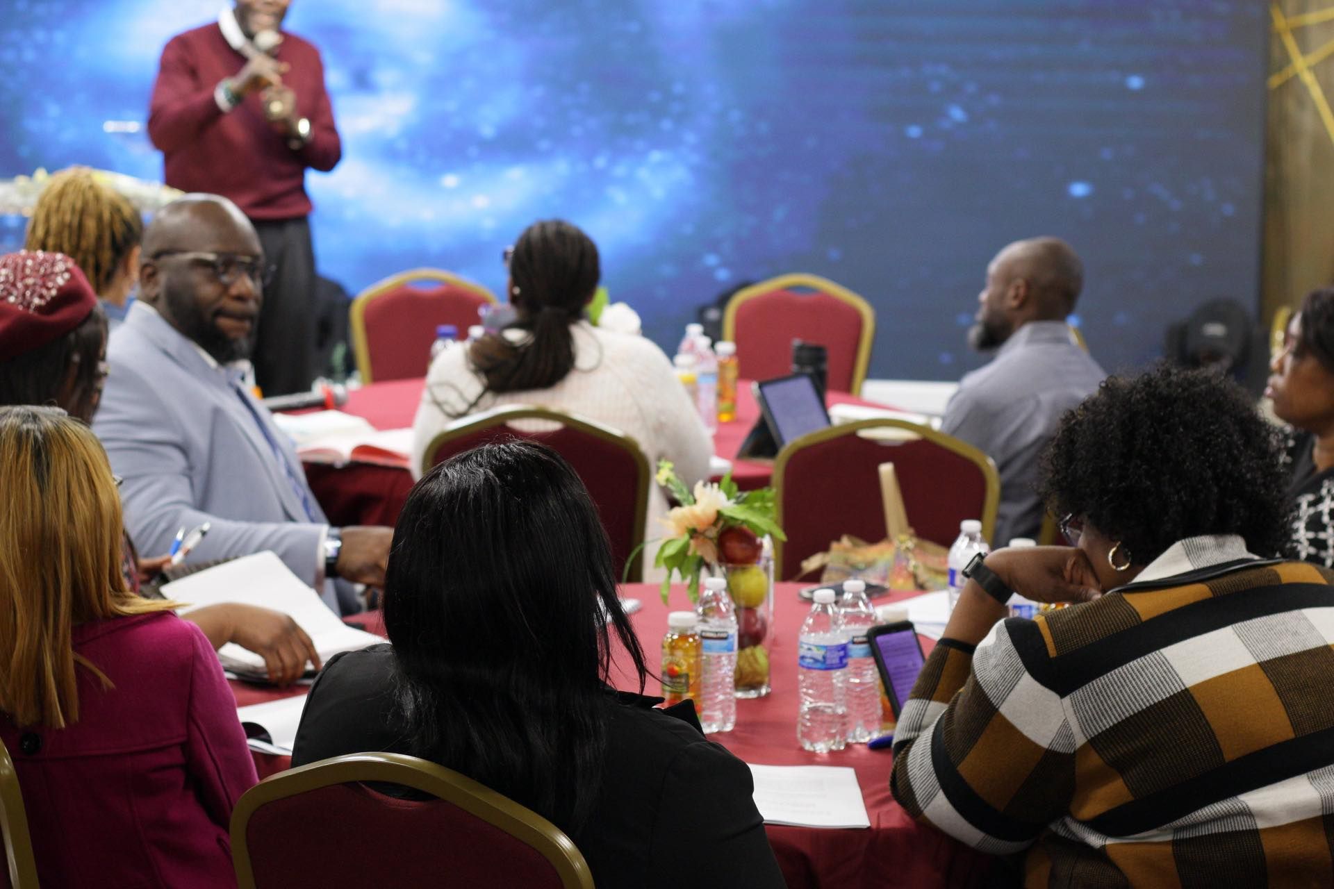 People at a conference table listen to a speaker. The room has a screen with a galaxy backdrop.
