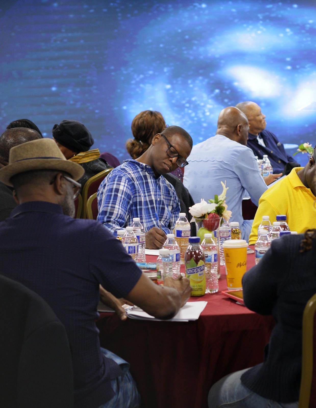 People seated at a table, some writing or looking down, in a room with a blue background.
