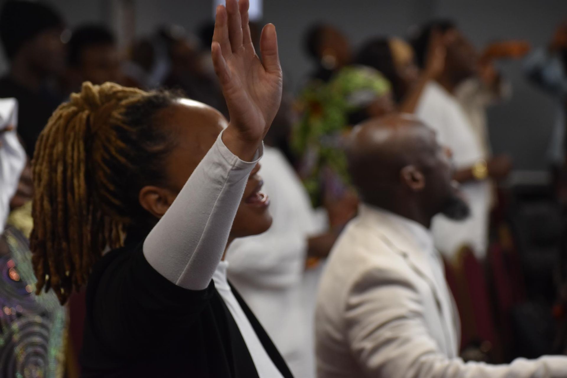Woman with dreadlocks raises hand in worship during a church service, others in background.