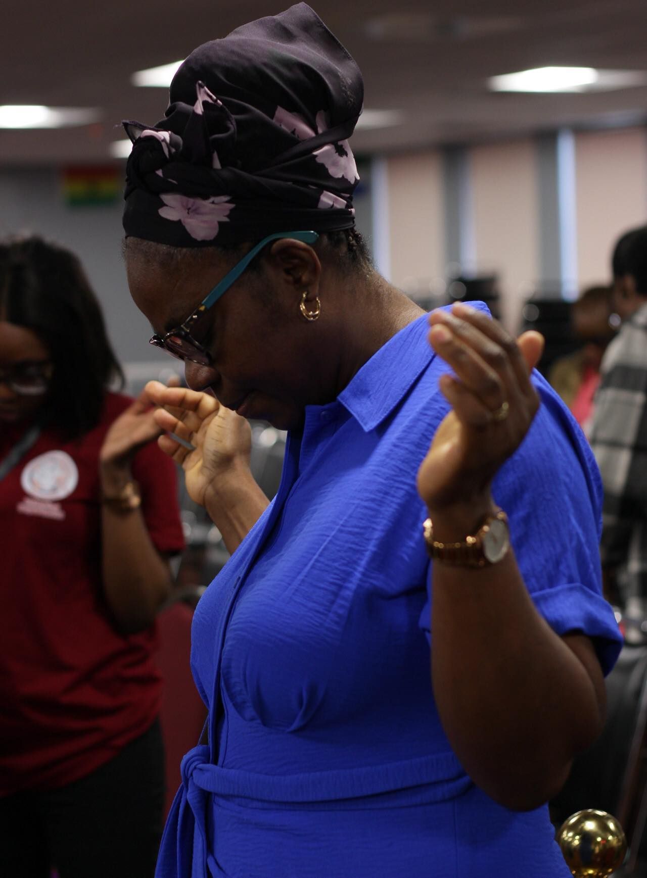 Woman in blue dress with headwrap praying, hands raised, in a room with others.