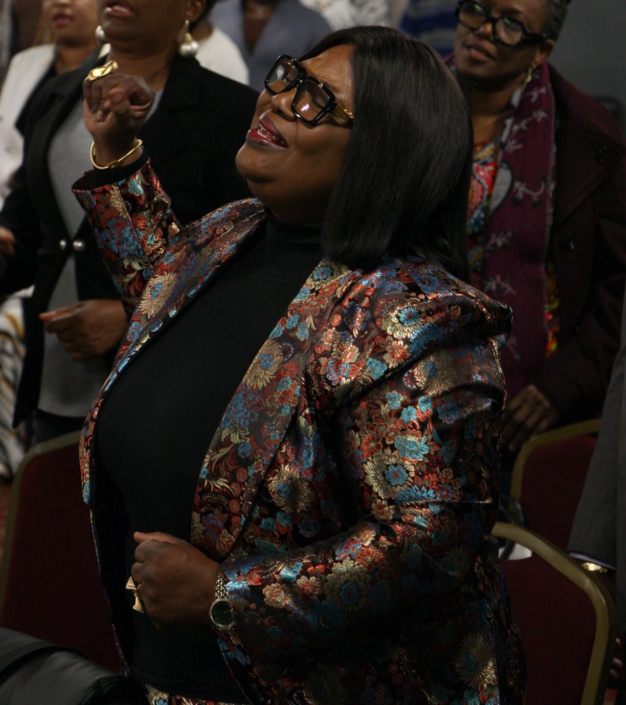 Woman in patterned blazer, raised arm, looking up, possibly in church.