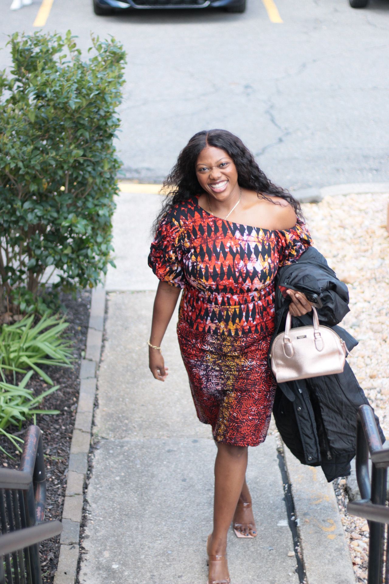 Woman in colorful off-shoulder dress smiling, walking up outdoor stairs, holding bag and jacket.