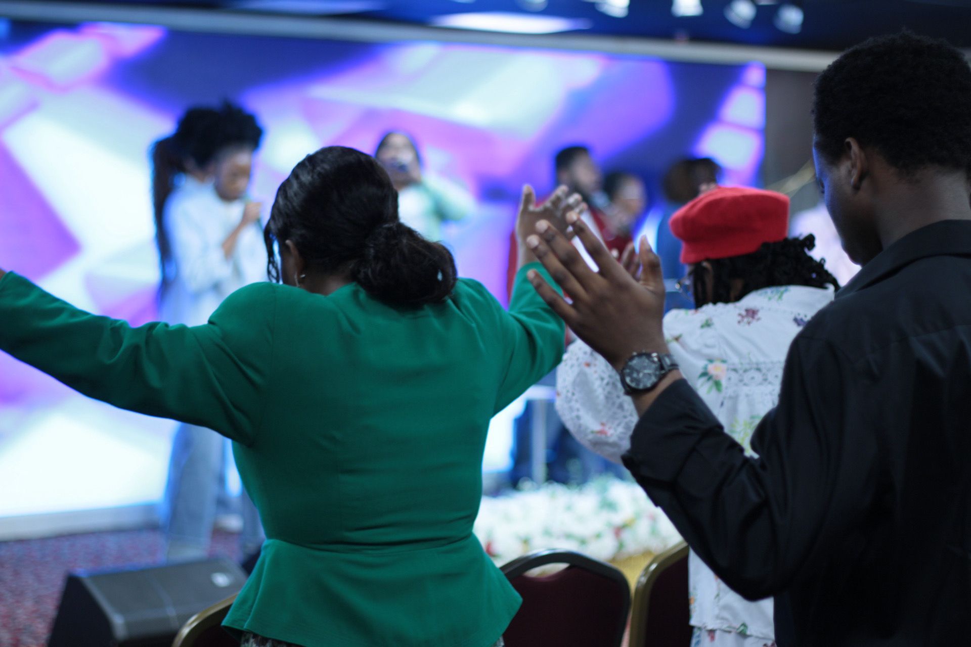 People in a church service; woman in green jacket raises hands, others clap, stage in background.