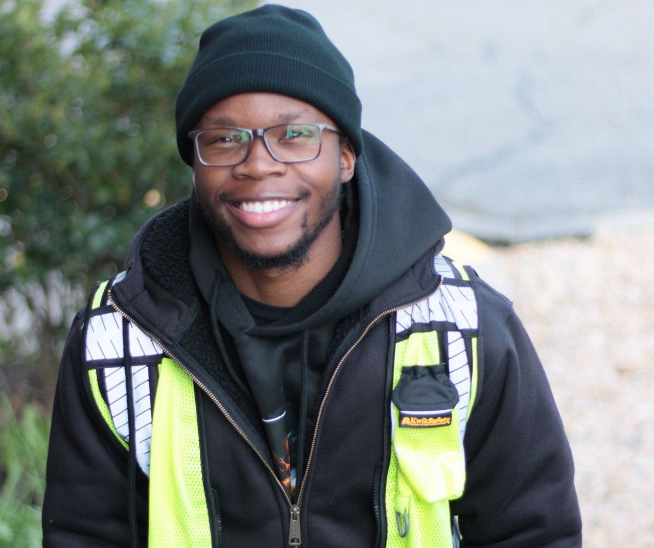 Man wearing glasses, black beanie, and reflective vest smiles outdoors.