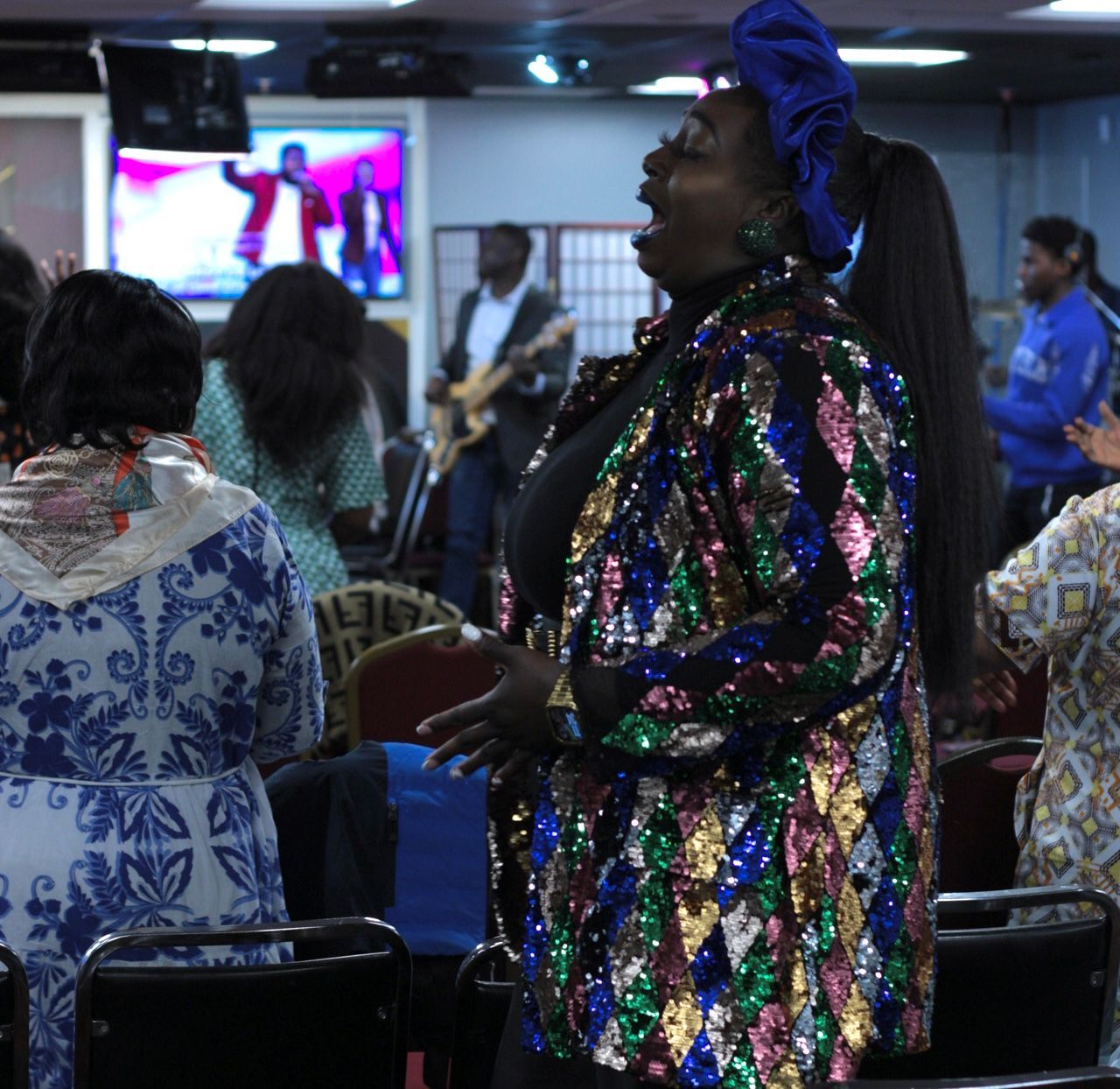 Woman singing in a church, wearing a sequined jacket and blue headscarf, with others in background.